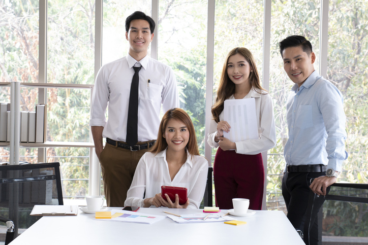 Group of young Asian business people working and communicating while at the office desk together with colleagues. Business people discussing over new business project in office. Group of business workers smiling happy.